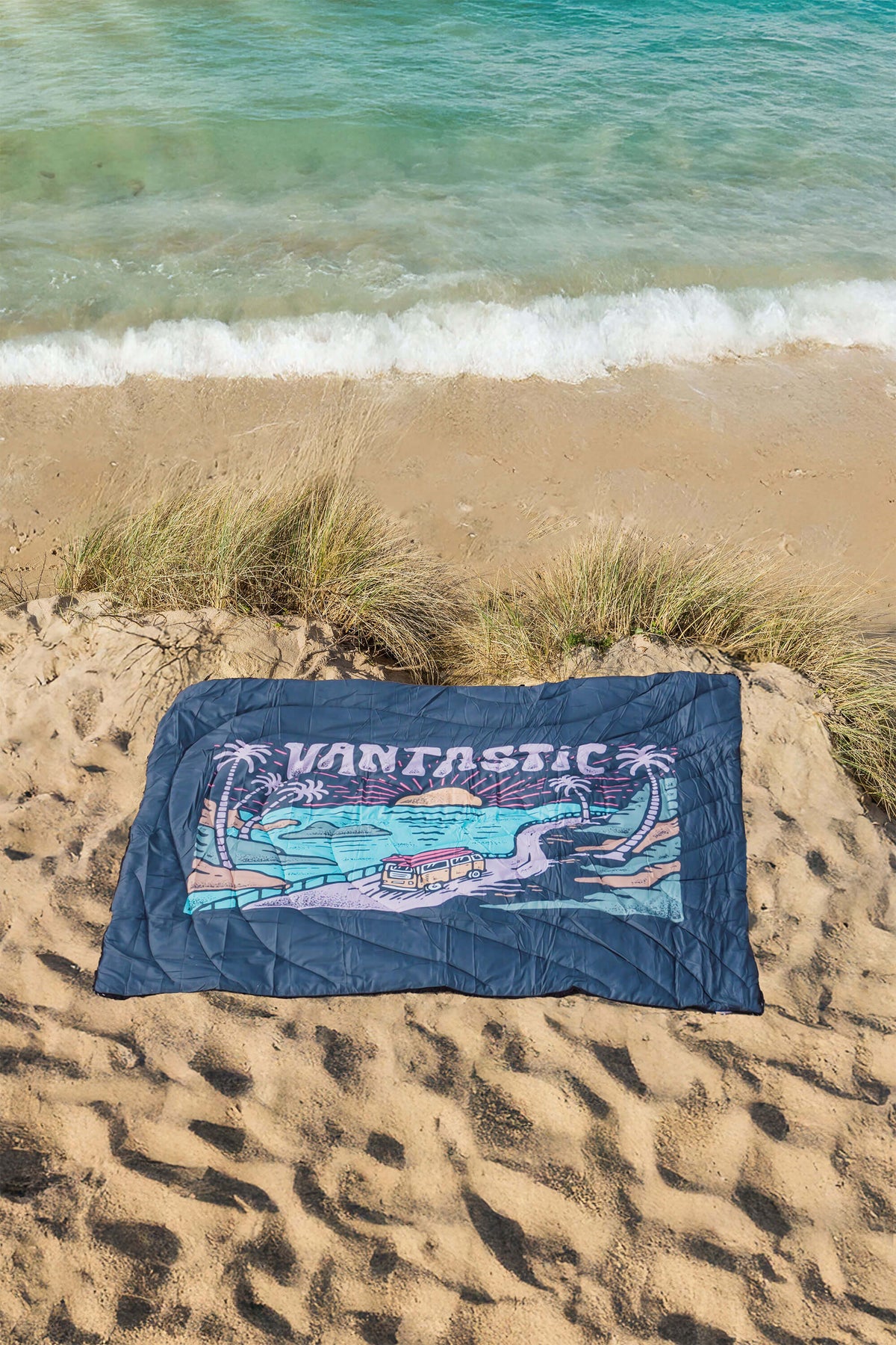 Navy blue camping blanket with 'Vantastic' design on a sandy beach with ocean waves.